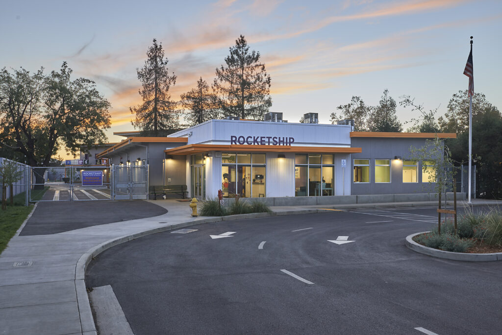 Rocketship school entrance at dusk, showcasing the modern building and lit windows, with trees and an American flag.