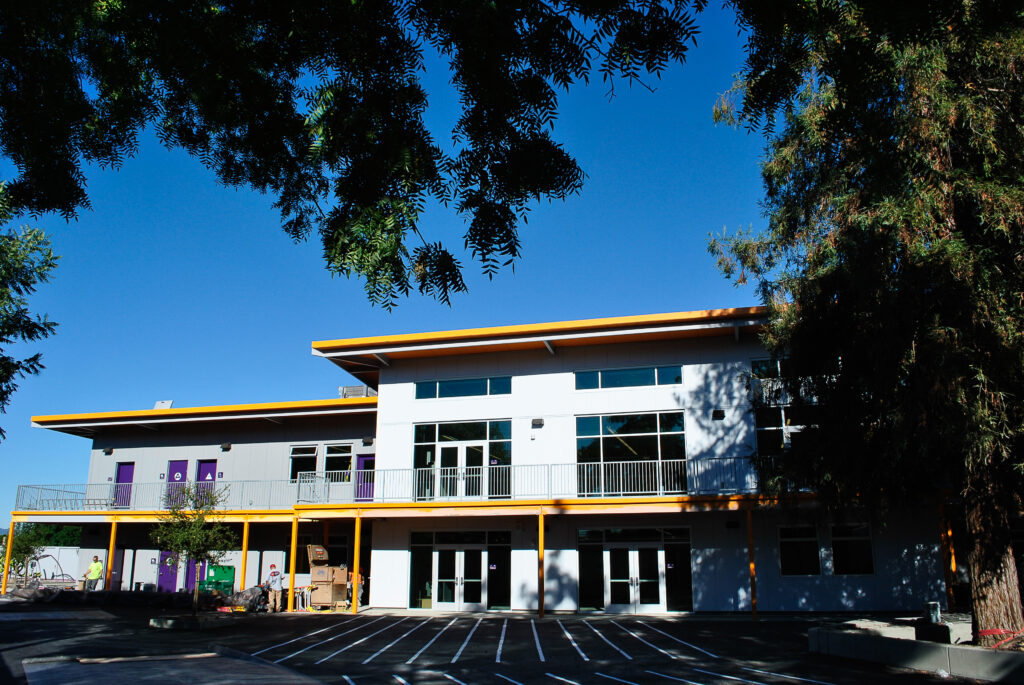 Rocketship school building, partially covered by tree foliage, with students and construction equipment in view.