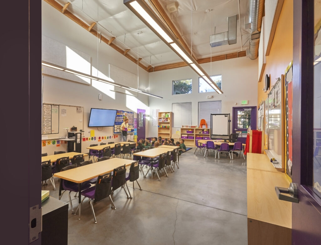 Brightly lit kindergarten classroom at Rocketship, featuring desks, chairs, and a teacher conducting a lesson with students.