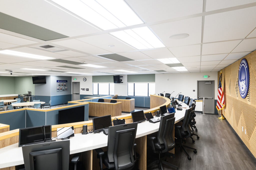 A wide view of a modern school district board room featuring a large curved white desk with black chairs and monitors, a wooden accent wall with a large blue district logo, and an American flag.