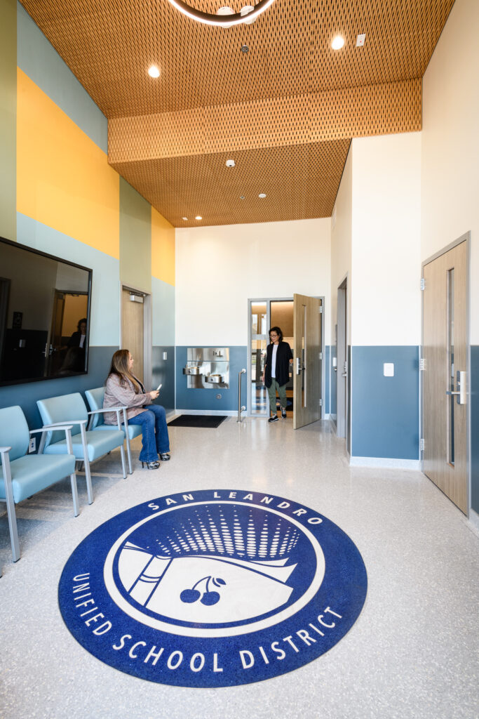 A tall, modern office lobby with a large blue circular "San Leandro Unified School District" logo rug on the floor. The room features light blue chairs, a drinking fountain station, and a high ceiling with wood-slat acoustic panels and a halo-style light fixture.
