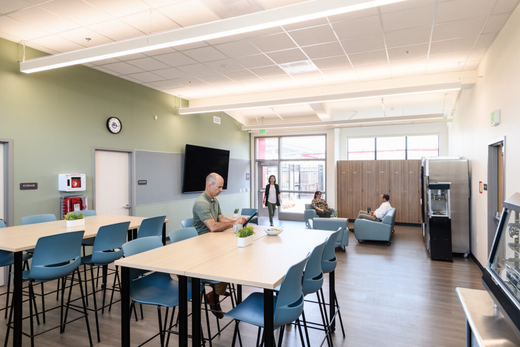 Interior of a modern school district staff room with long light-wood tables, blue high-back chairs, and a lounge area with soft seating. A man sits at a table using a tablet, while other staff members are in the background near wooden lockers and a wall-mounted TV.
