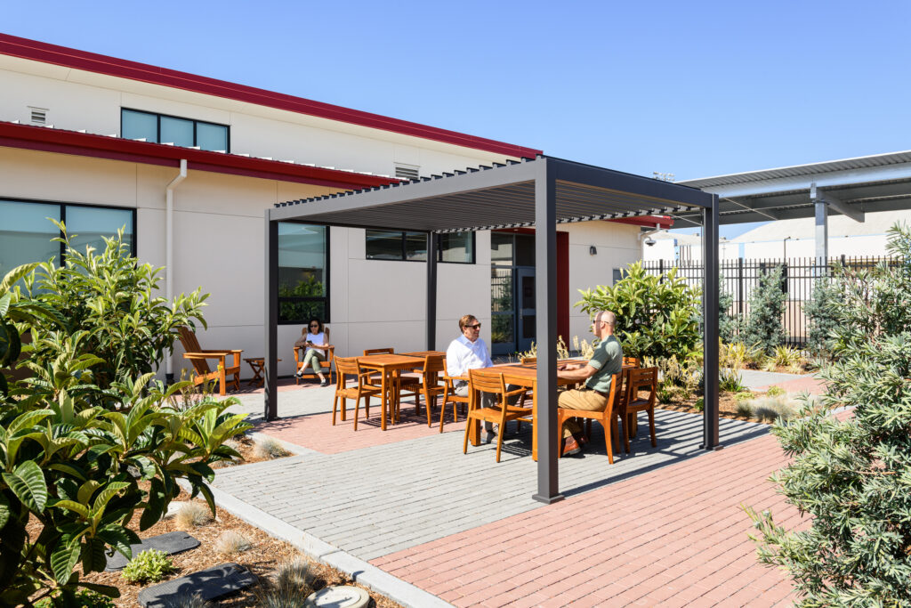 A grey modular office building with red roof trim, an external metal staircase, and a concrete courtyard with small trees under a clear blue sky.