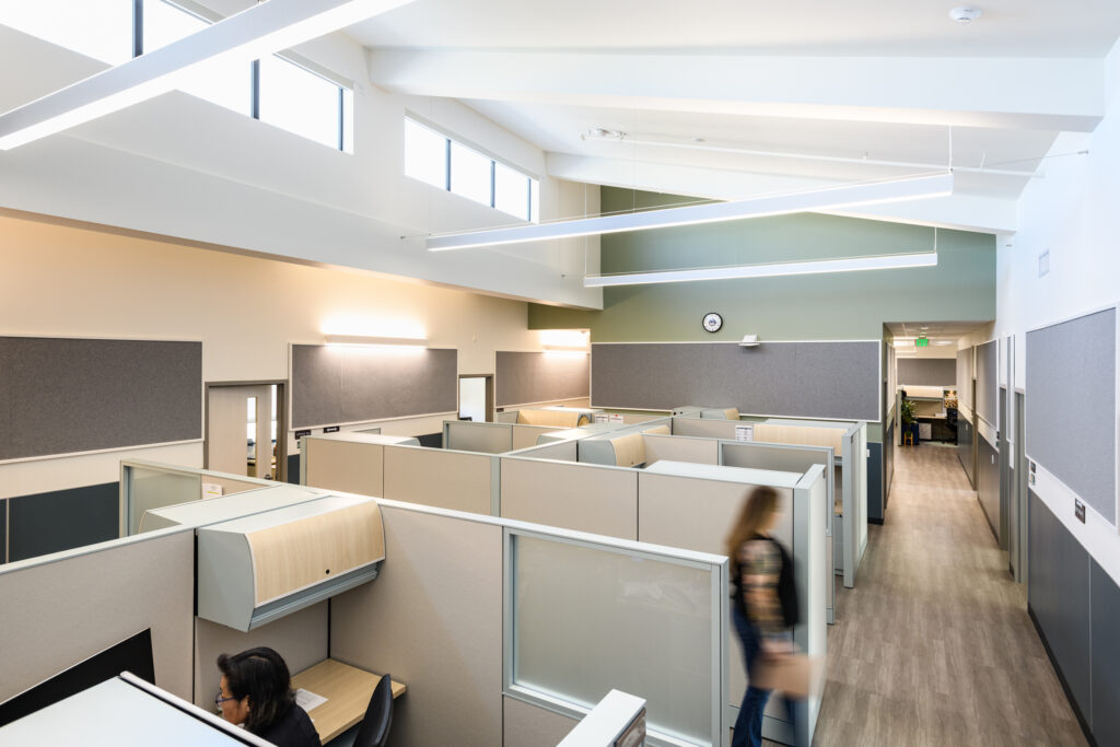 Interior of a modern office with tall white vaulted ceilings, light-toned cubicles, wood-look flooring, and a person walking down a wide hallway.
