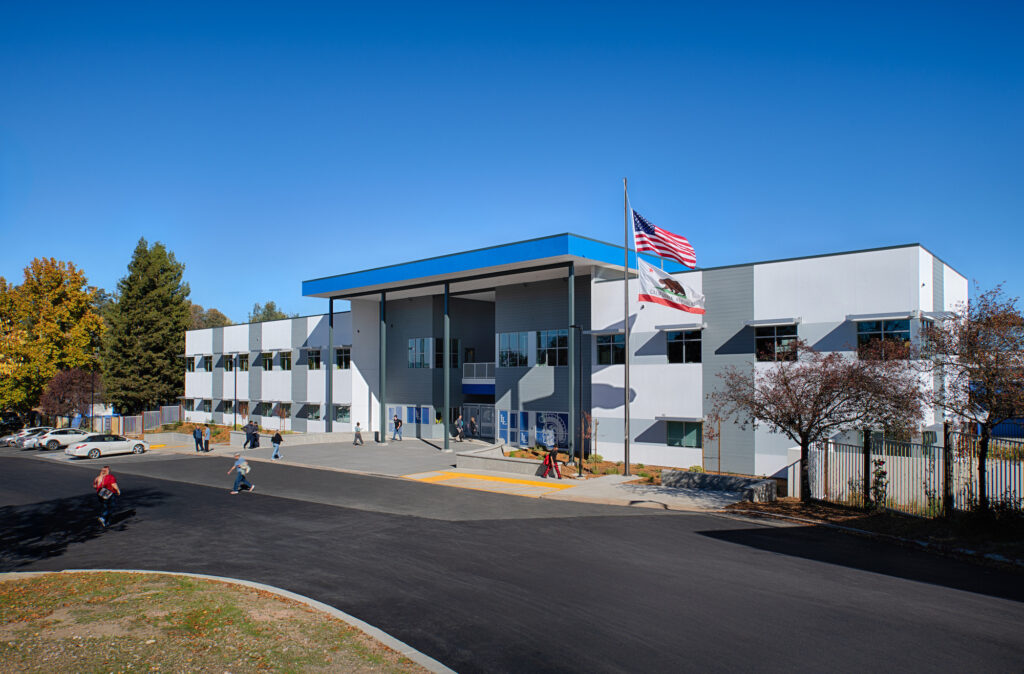 A wide exterior view of a modern two-story white and grey building with blue roof accents. The entrance features tall pillars, glass windows with "LL" branding, and two flagpoles flying the American and California flags.