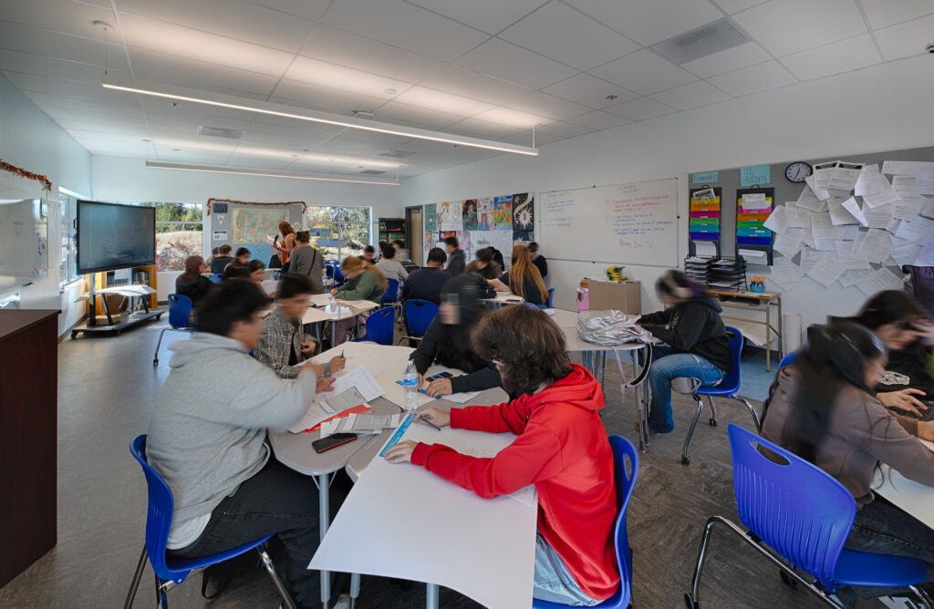 A vibrant, well-lit classroom filled with students working at white modular desks with blue chairs. The room features a large digital display, whiteboards covered in lesson notes, and modern linear LED lighting under a drop ceiling.