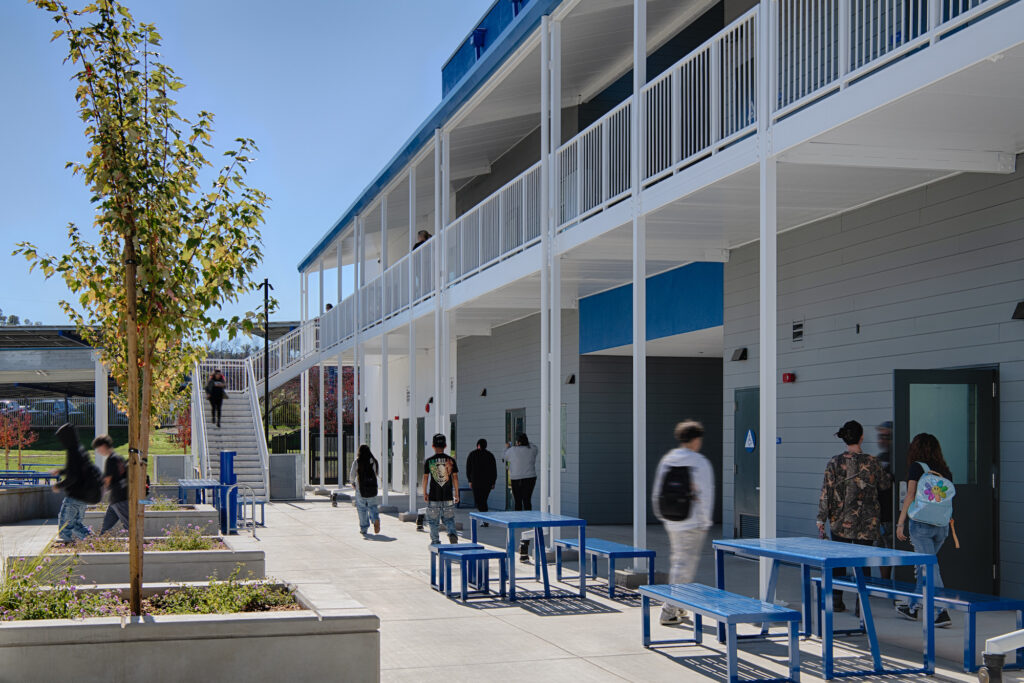 Exterior view of a modern two-story grey modular school building with white railings and stairs. Students walk through a concrete courtyard with blue metal picnic tables and landscaped planters under a clear blue sky.