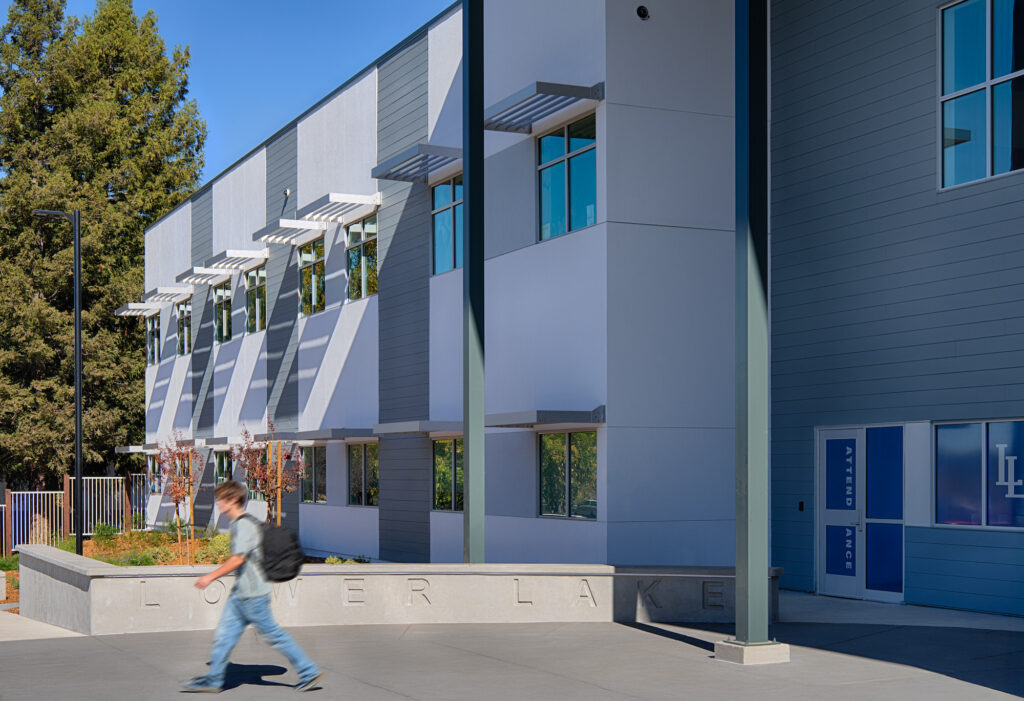 A student with a backpack walks in front of a modern two-story school building. The building features grey and white siding, large windows with white awnings, and a prominent blue "ATTENDANCE" sign and "LOWER LAKE" lettering on a concrete wall. A large tree is on the left under a clear blue sky.