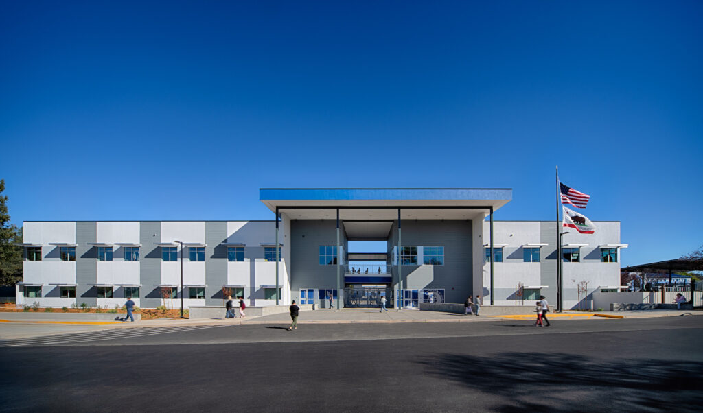 A wide-angle front view of a large, modern two-story school building with grey and white siding, a tall central covered entrance, and a flagpole flying the American and California flags.