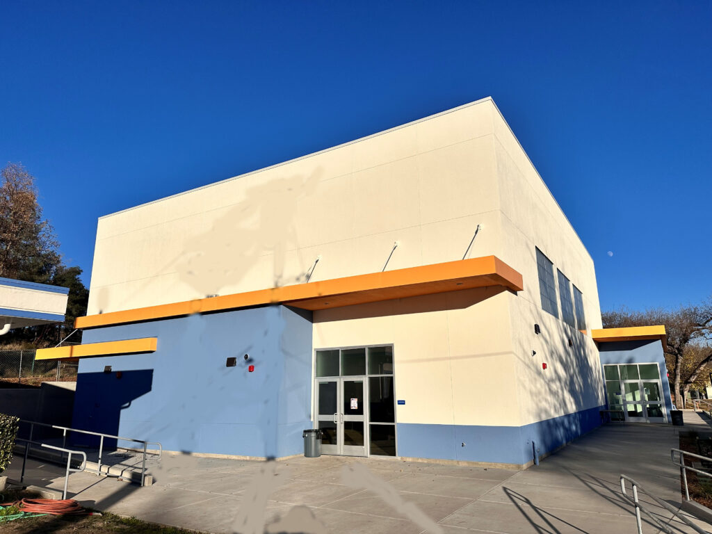 Exterior of a modern gymnasium with a cream and blue facade, featuring bold orange overhangs, large windows, and a professional concrete entryway under a clear blue sky.