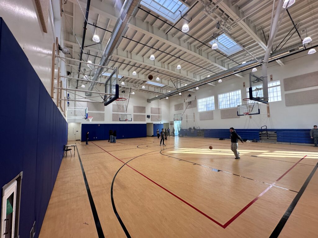 Interior view of a well-lit gymnasium with a wooden basketball court where several people are playing. The gym has high ceilings with exposed beams and skylights, multiple retractable basketball hoops, blue wall padding, and blue bleachers along the back wall.
