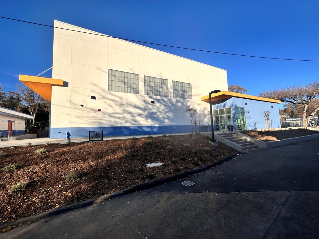 A side view of a large, modern gymnasium building. It has a cream-colored upper section, a blue lower section, and a bright orange overhang above a side entrance with concrete stairs. Three large, square translucent panels are high on the wall. The foreground features landscaping with dark wood chips and small plants under a clear blue sky.