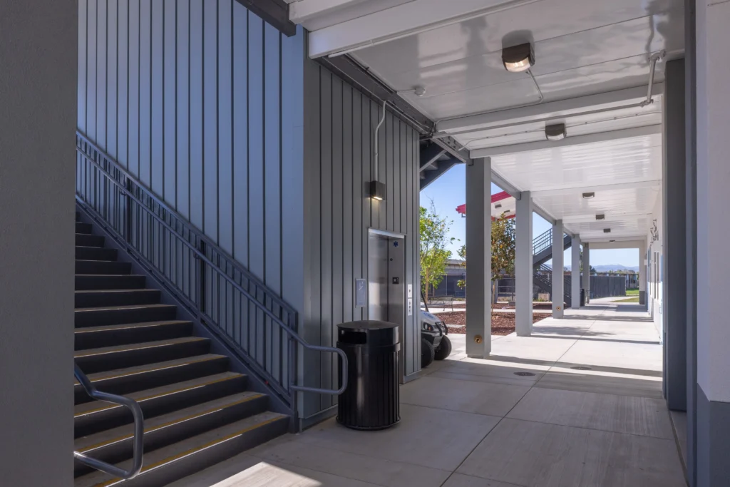 Architectural view of a school's exterior featuring a grey staircase, elevator access, and a long covered walkway.