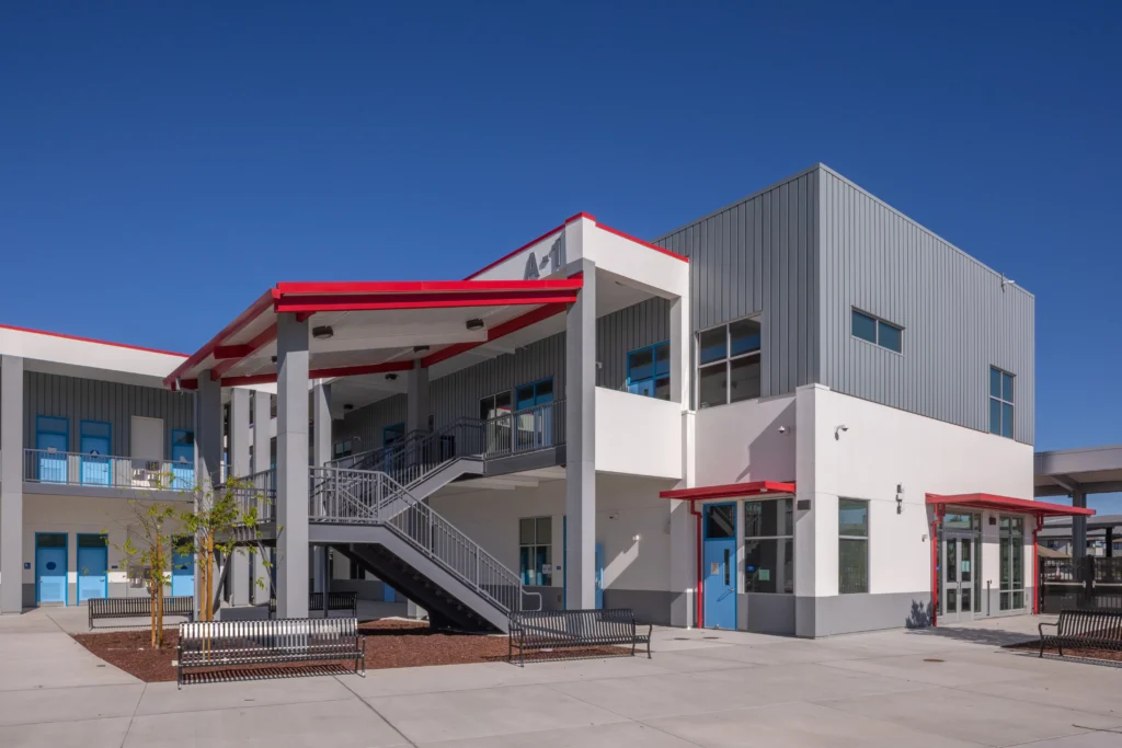 Exterior view of a two-story school building with grey siding, red roof accents, blue doors, and an outdoor staircase.