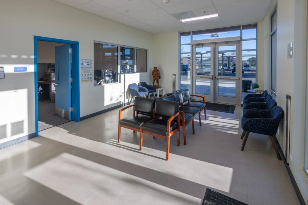 Interior of a school waiting room with black and blue chairs, an open blue-framed office door, and bright sunlight.