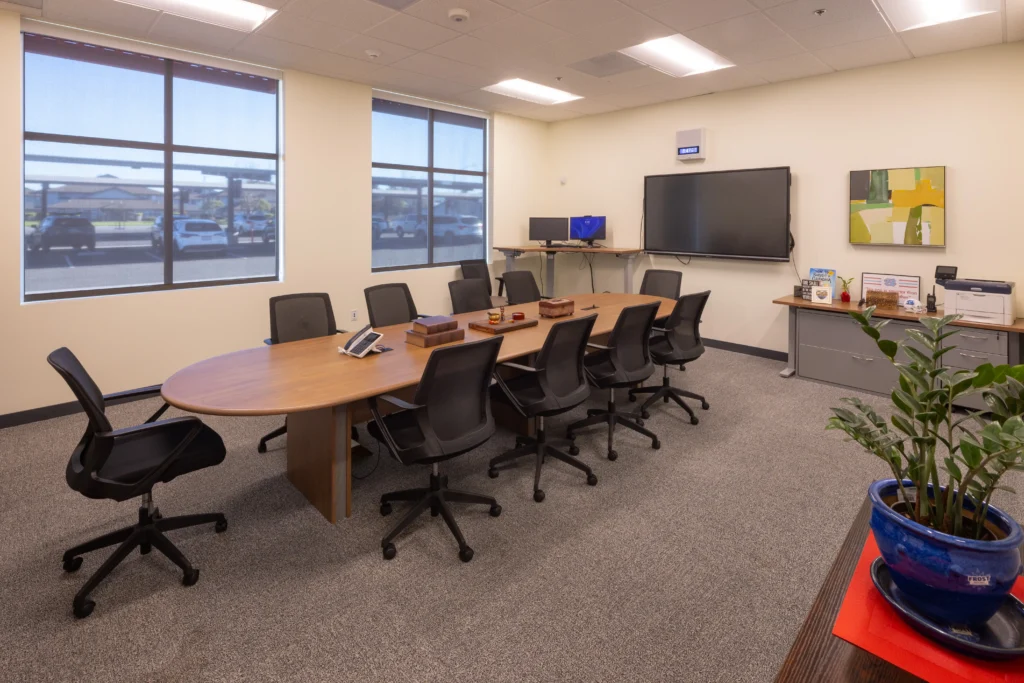 A modern school conference room with a large oval wooden table, black office chairs, and a wall-mounted display.